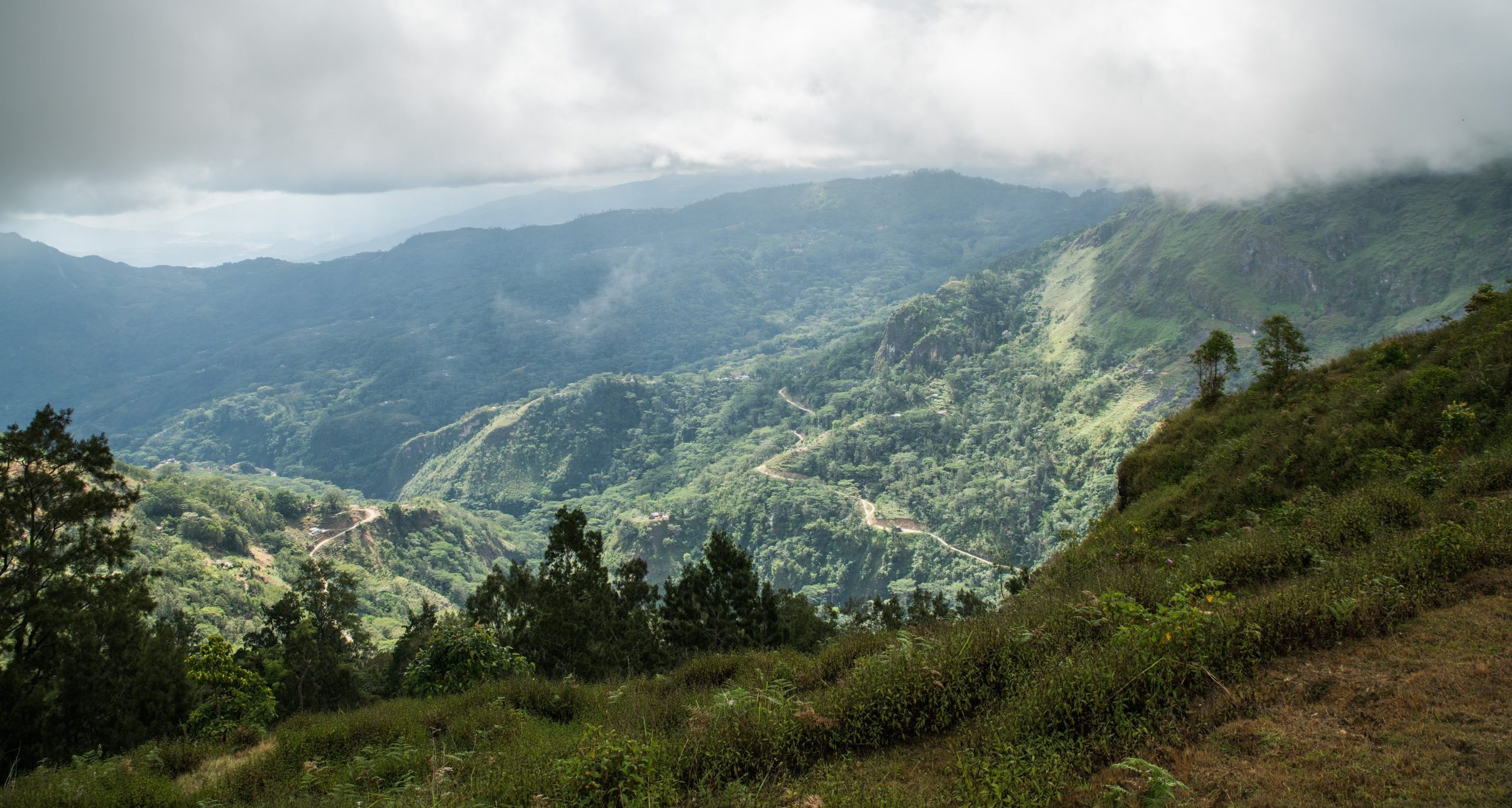 Image shows high altitude region of Emera in Timor Leste. An expanse of grey cloud cover reaches across several mountains with shrubs and trees in close by.